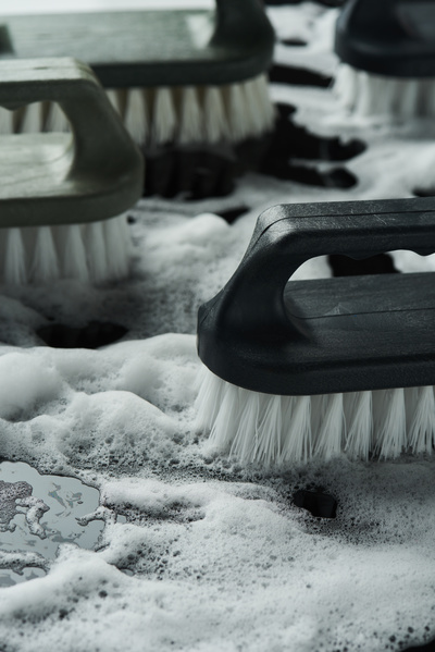 Close-up of a household iron brush with a black handle and white bristles in soap foam against the background of other cleaning brushes